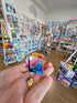 Hand holding Motus Mini CMY Cube inside gift shop with toys and travel items, colourful fidget toy Adventure Awaits