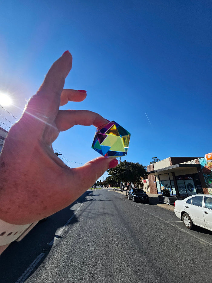 Hand holding Motus Mini CMY Cube against blue sky on country town street, colourful travel fidget toy Adventure Awaits