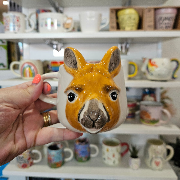Hand holding a ceramic kangaroo-shaped mug with a blurred background of shelves with more mugs.