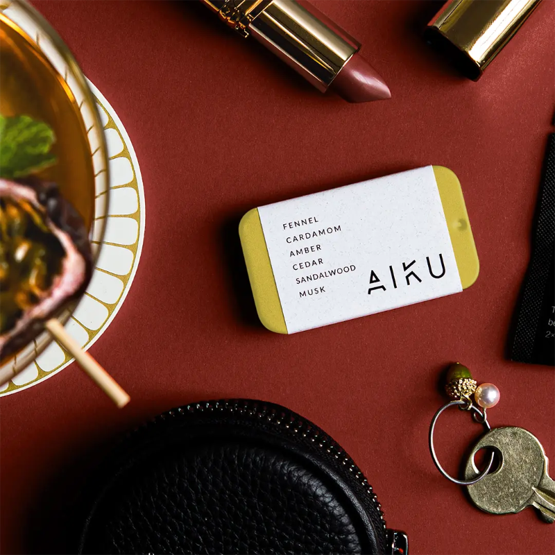 AIKU tin of solid perfume in Ember with woody fragrance notes beside lipstick, keys and handbag — Adventure Awaits