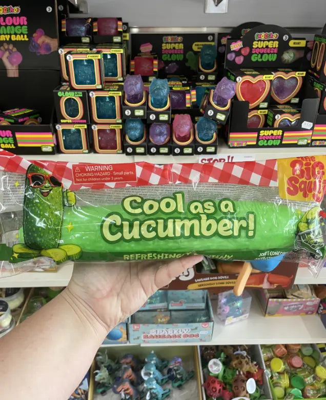 Hand holding a giant green Cool as a Cucumber squishy toy in retail packaging — Adventure Awaits