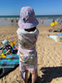 Child on a beach wearing a colorful hat and towel, with ocean and people in the background.