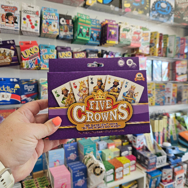 Hand holding a 'Five Crowns' card game box in a store aisle with various games.