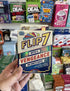Flip 7 With a Vengeance card game pack held in store with colourful game shelf Adventure Awaits