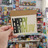 Hand holding a 'Happy Birthday' card in front of a store shelf with various items.
