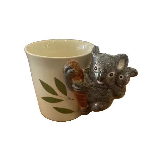 Cream ceramic koala mug with sculpted koala and joey handle beside eucalyptus leaves — Adventure Awaits