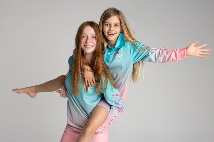 Two girls smiling in aqua and pink UPF50+ fishing dresses, one giving the other a piggyback — Adventure Awaits