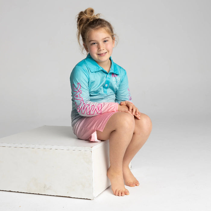 Young girl wearing a turquoise long-sleeve collared top with pink shorts while seated — Adventure Awaits