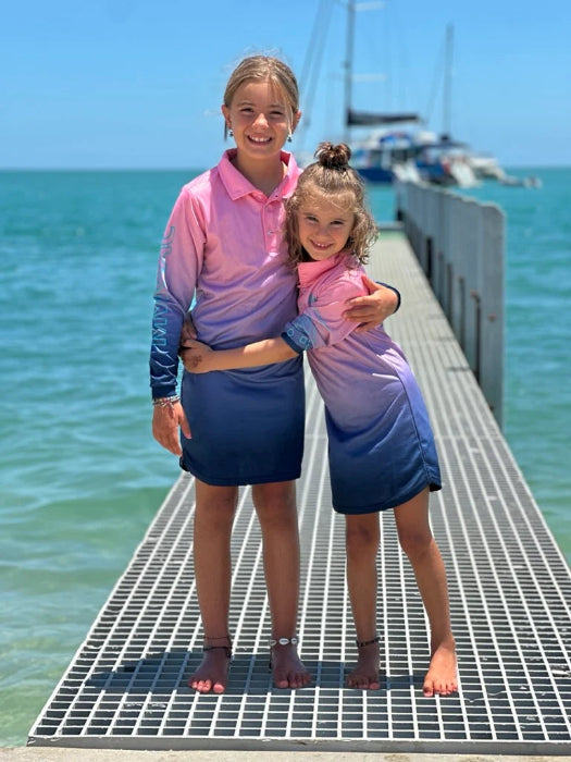 Two girls hugging on a jetty in matching pink and blue UPF50+ fishing dresses by the sea — Adventure Awaits