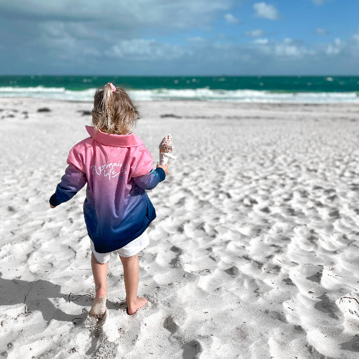 Girl walking barefoot on a beach in a pink and blue Mermaid Life fishing shirt, holding an ice cream — Adventure Awaits