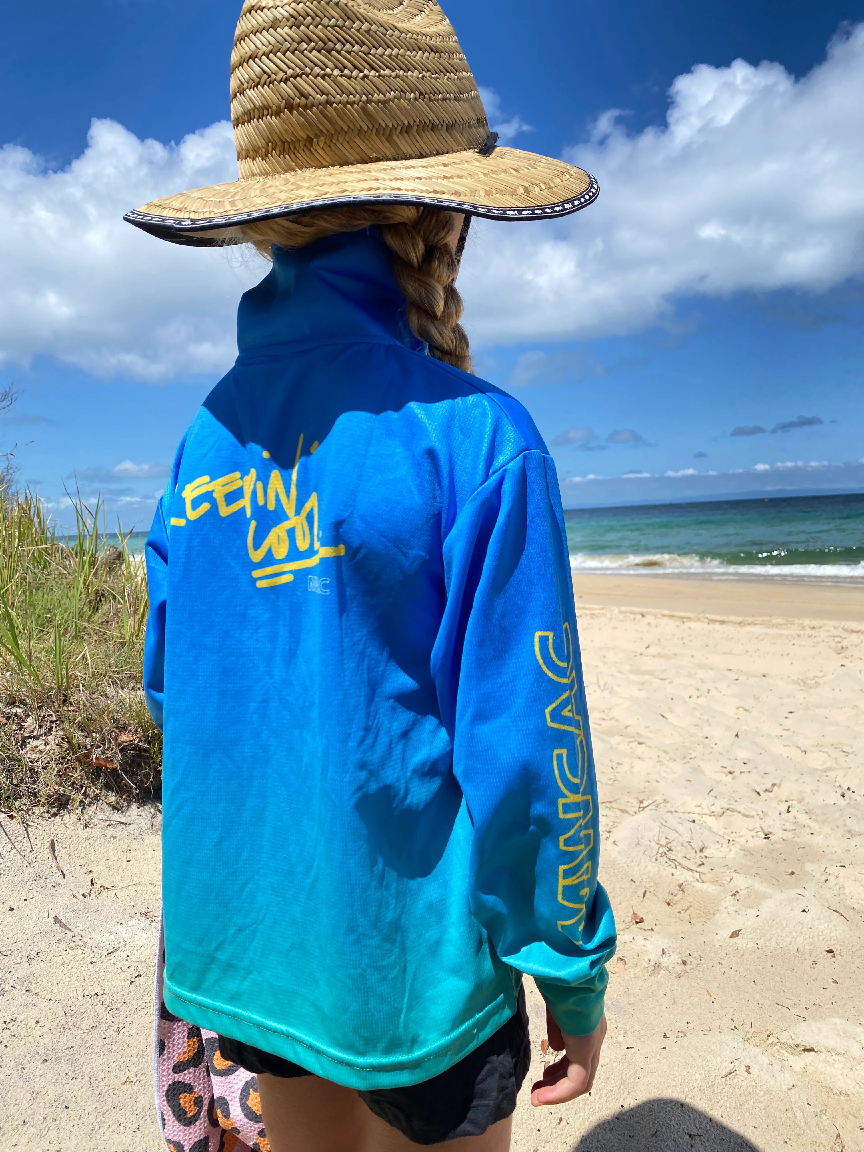 Back view of girl in a blue and aqua long-sleeve fishing shirt and straw hat walking by the beach — Adventure Awaits