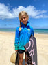 Girl standing on a sunny beach in a blue and aqua long-sleeve fishing shirt holding a towel — Adventure Awaits