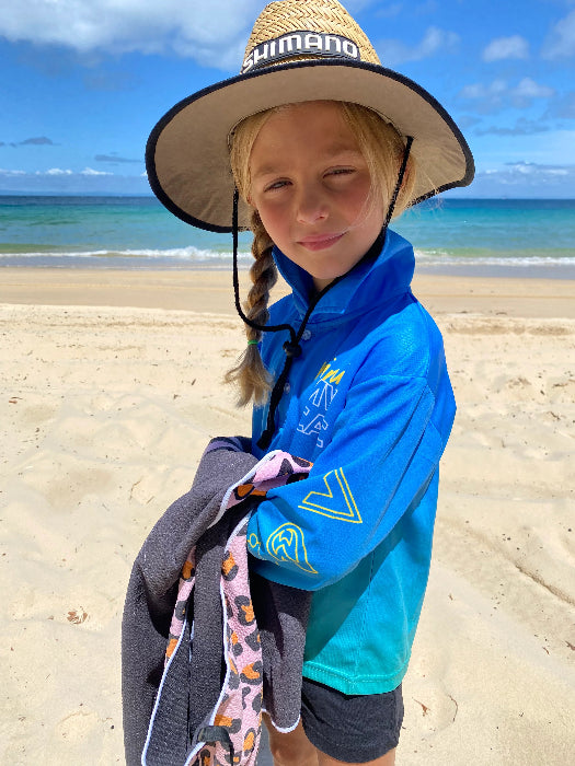 Girl in a blue long-sleeve fishing shirt and wide-brim hat standing on a sunny beach — Adventure Awaits