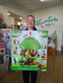 A person holding up the retail packaging box for the 'Pop L'arbre magique des Klorofils / The Klorofil Magic Tree' toy set in a toy store. The box features a green treehouse design with various colorful accessories.