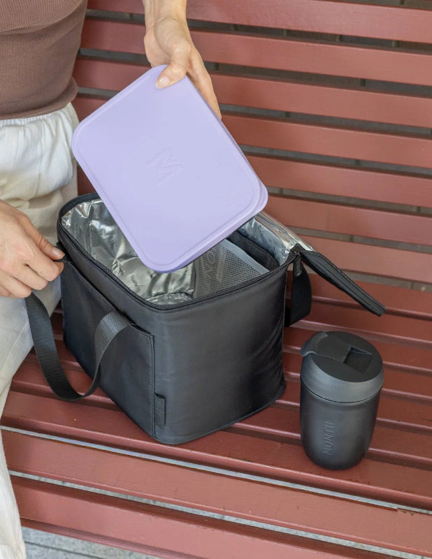 Person placing a purple lunchbox into an open black insulated lunch bag beside a travel cup — Adventure Awaits