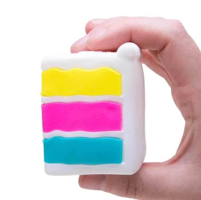 Hand holds a NeeDoh Piece O’ Cake squishy toy showing white icing with yellow, pink and blue layers — Adventure Awaits