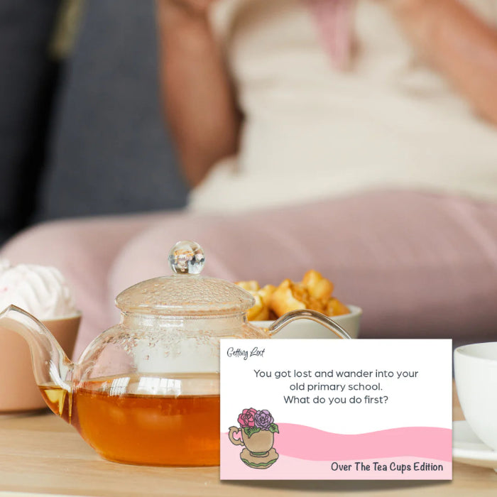 Glass teapot and Getting Lost Over The Tea Cups Edition card on a relaxed tea table — Adventure Awaits