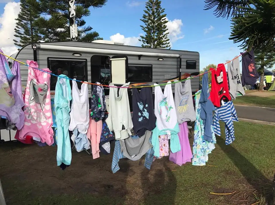 Children's clothing hanging on a pegless line outside a caravan with trees and blue sky in the background.