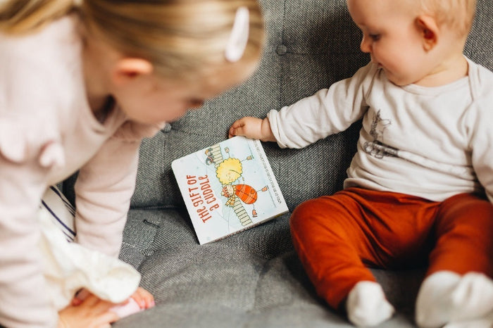 Child sharing The Gift of a Cuddle board book with baby on grey couch — Adventure Awaits