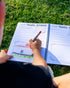 Colourful picture being drawn into the kids travel journal on the grass by young child 