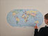 A person pointing at a large oval-shaped world map sticker mounted on a plain wall - Adventure Awaits