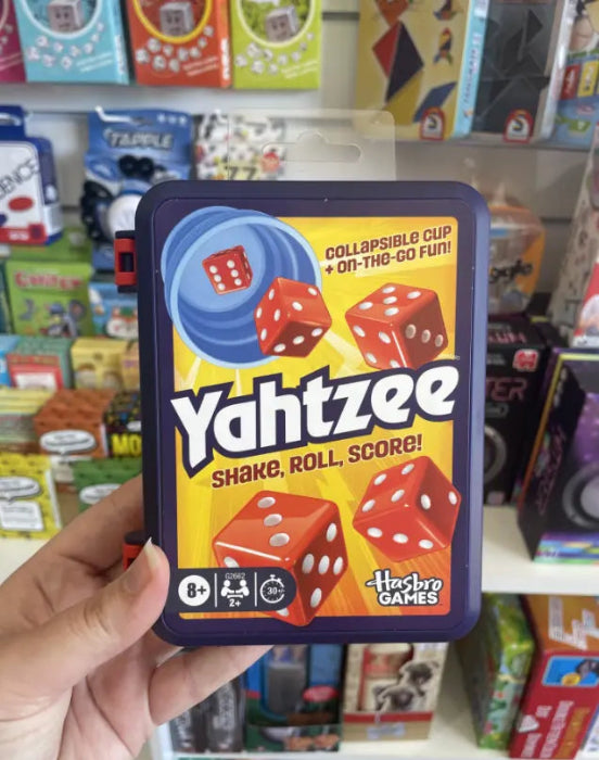 Hand holding Yahtzee travel game case with red dice design in front of a colourful games display — Adventure Awaits