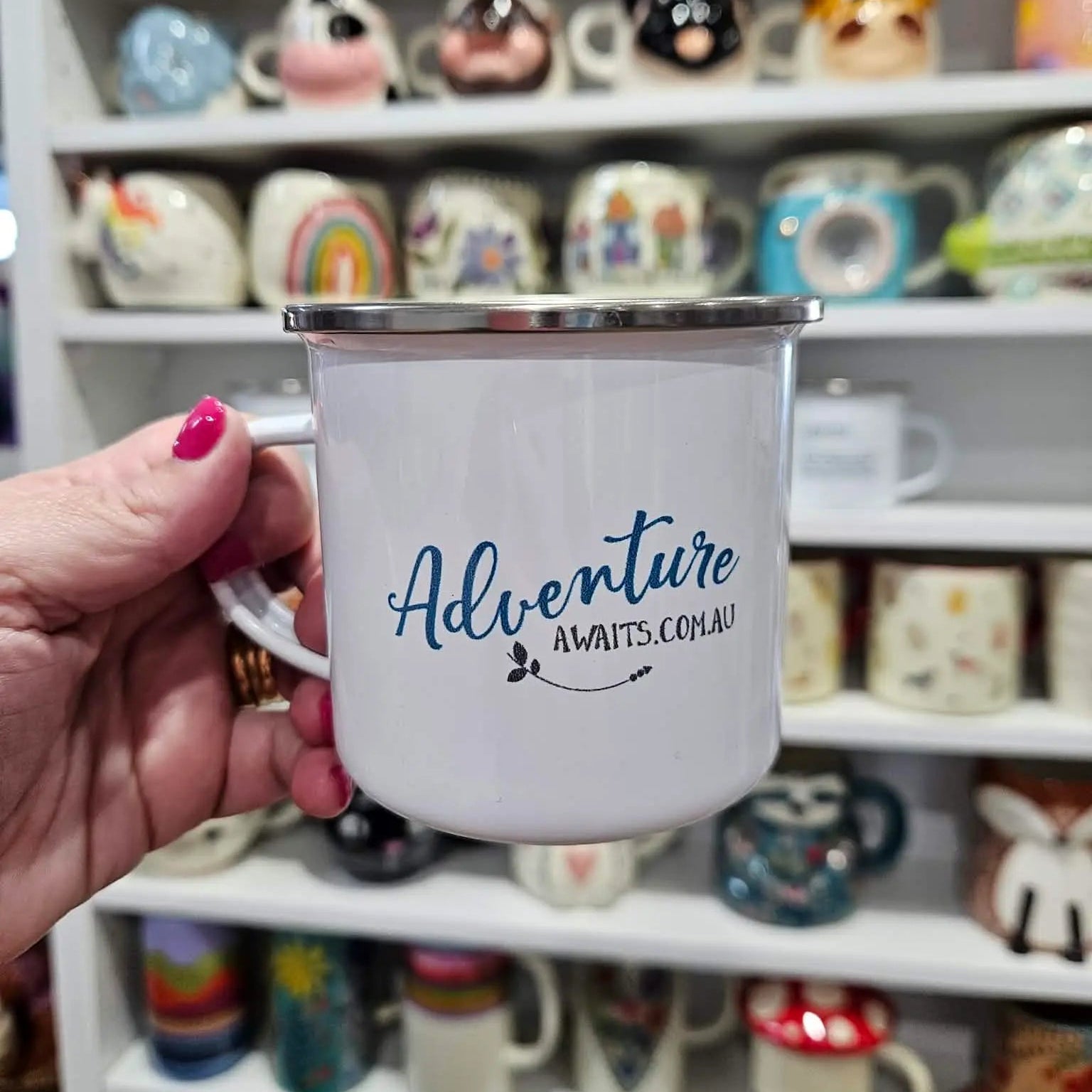 White mug with 'Adventure Awaits.com.au' text held in front of a shelf of various mugs.