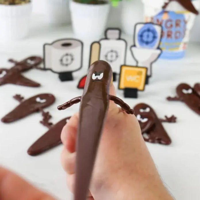 Hand holding a brown Angry Turds flying poop game piece with toilet targets in background — Adventure Awaits