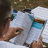 Camper reading Outback Queensland guide in Big Lap Bible beside enamel mug at campsite, road trip travel guide Adventure Awaits
