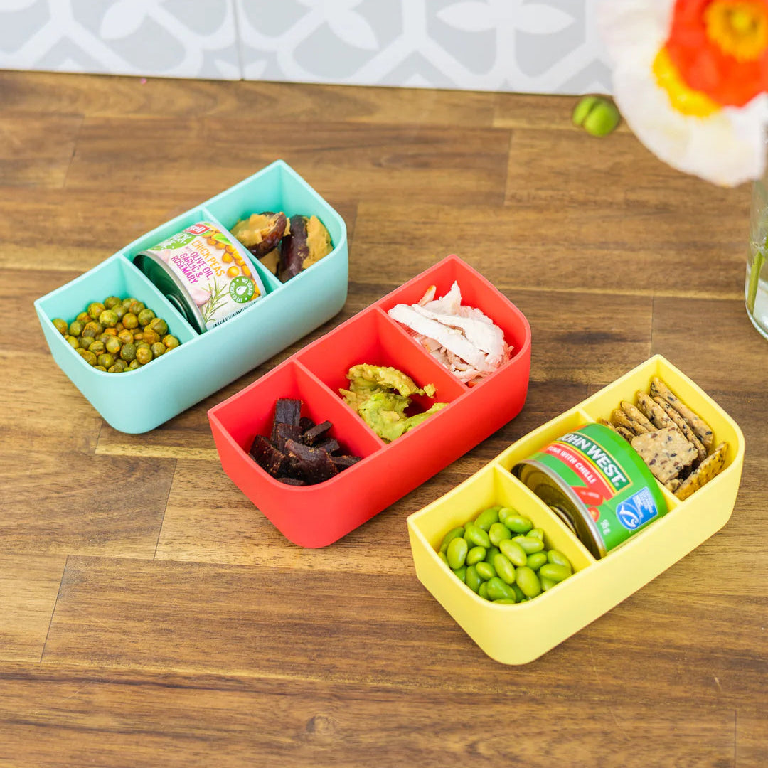 Three colorful bento boxes with food on a wooden surface, with flowers in the background.