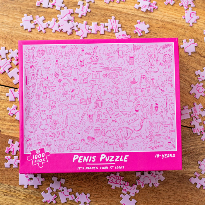 Pink 1000-piece Penis Puzzle box surrounded by matching jigsaw pieces with cheeky illustrated designs — Adventure Awaits