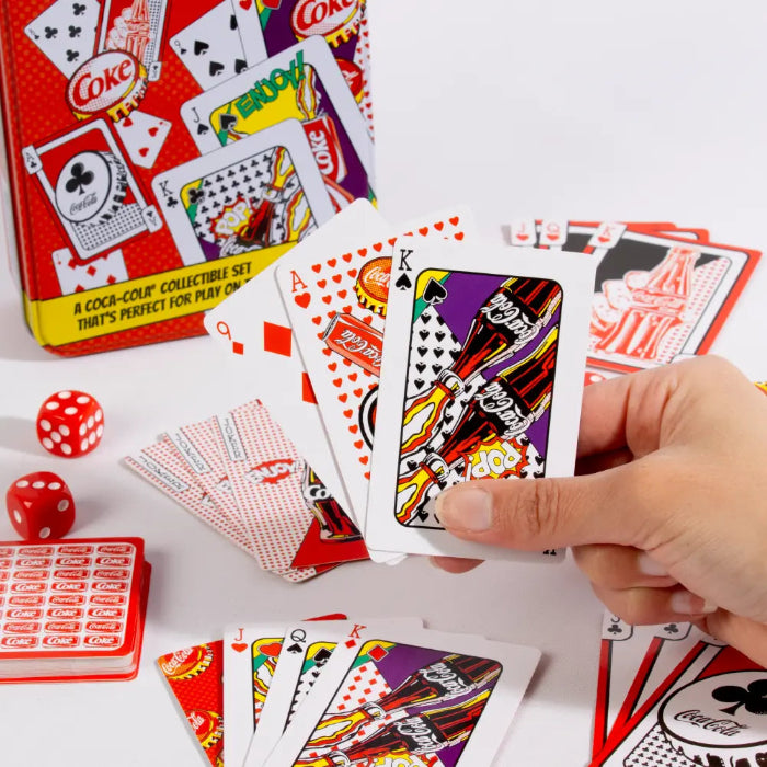 Hand holding Coca-Cola playing cards with dice and collectible tin — Adventure Awaits