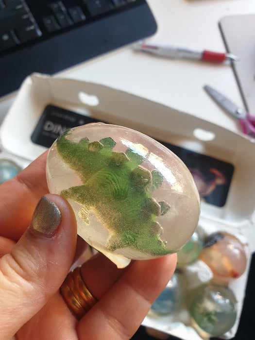 Hand holding clear dinosaur egg fragranced soap with green dinosaur figure inside — Adventure Awaits