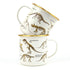 Two cream enamel dinosaur mugs stacked with gold rims and skeleton illustrations Adventure Awaits