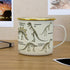 Cream enamel dinosaur mug with gold rim on desk, skeleton illustrations and study notes Adventure Awaits