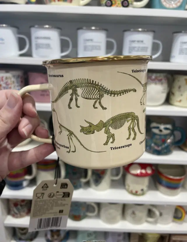 Cream enamel dinosaur mug held in store with Triceratops and Velociraptor skeletons Adventure Awaits