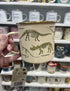Cream enamel dinosaur mug held in store with Triceratops and Velociraptor skeletons Adventure Awaits