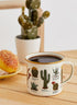 enamel mug with cactus illustrations filled with coffee on wooden table, gold rim camping mug styled with plant and pastry Adventure Awaits