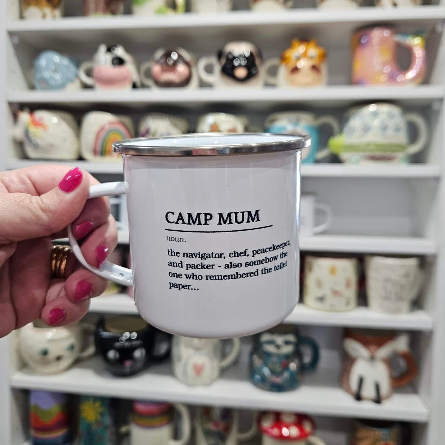 Person holding a 'Camp Mum' mug with a definition printed on it, in front of a shelf with various decorative mugs.