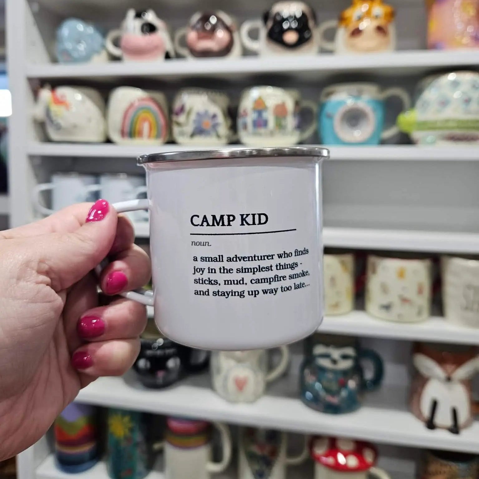Person holding a white mug with 'Camp Kid' definition text in front of a shelf of various mugs.