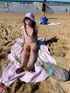 Child sitting on a beach with a floral swimsuit and sun hat, surrounded by beach items.