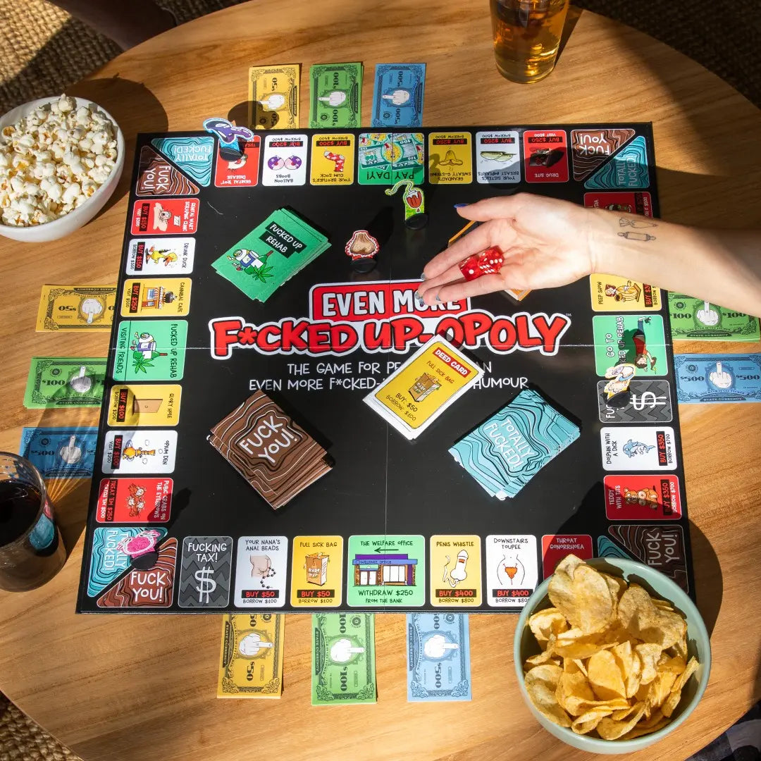 Board game 'Even More F**ked Up Opoly' on a table with snacks and drinks.