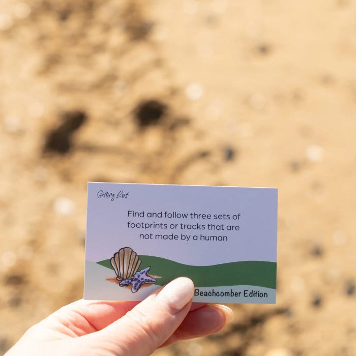 Hand holds a Getting Lost Beachcomber Edition card prompting players to follow animal tracks on the sand, Adventure Awaits