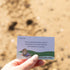 Hand holds a Getting Lost Beachcomber Edition card prompting players to follow animal tracks on the sand, Adventure Awaits
