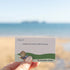 Hand holds a Getting Lost Beachcomber Edition card inviting one worry to be left with the tide, Adventure Awaits