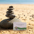 Stone tower beside a Getting Lost Beachcomber Edition card on a sandy beach, Adventure Awaits