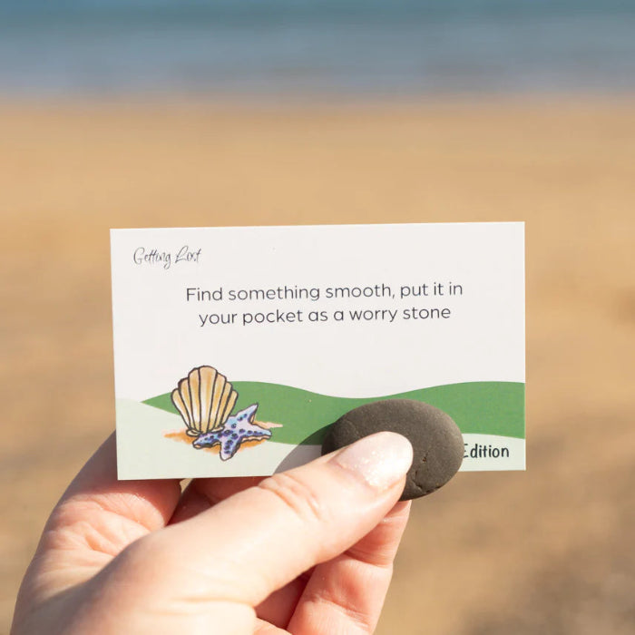 Hand holds a smooth worry stone beside a Getting Lost Beachcomber Edition card at the beach, Adventure Awaits