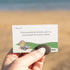 Hand holds a smooth worry stone beside a Getting Lost Beachcomber Edition card at the beach, Adventure Awaits