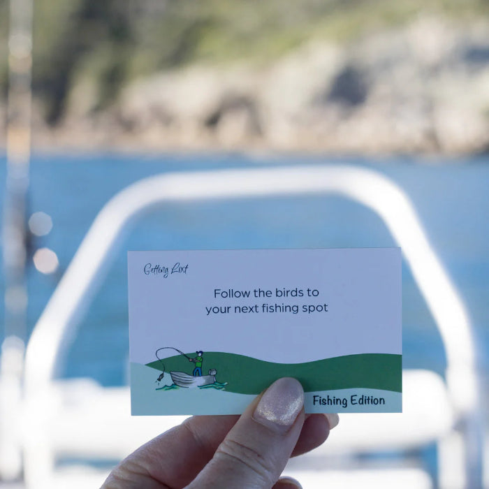 Hand holds a Getting Lost Fishing Edition card prompting anglers to follow birds to the next fishing spot, Adventure Awaits