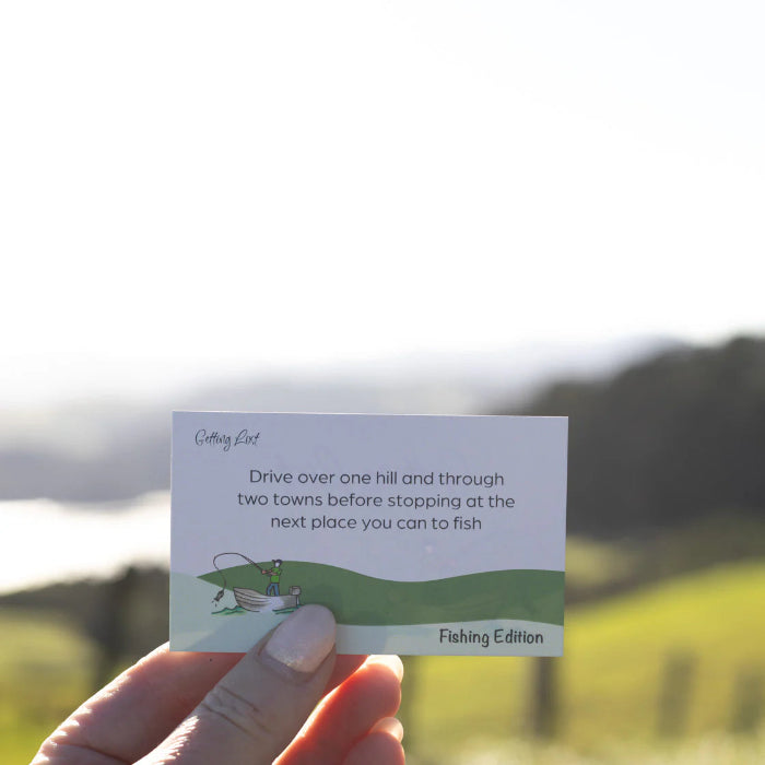 Hand holds a Getting Lost Fishing Edition card directing anglers over hills and towns to a fishing spot, Adventure Awaits